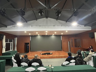 Lying on a low stage at the front of a seminar hall; audience members hold cameras